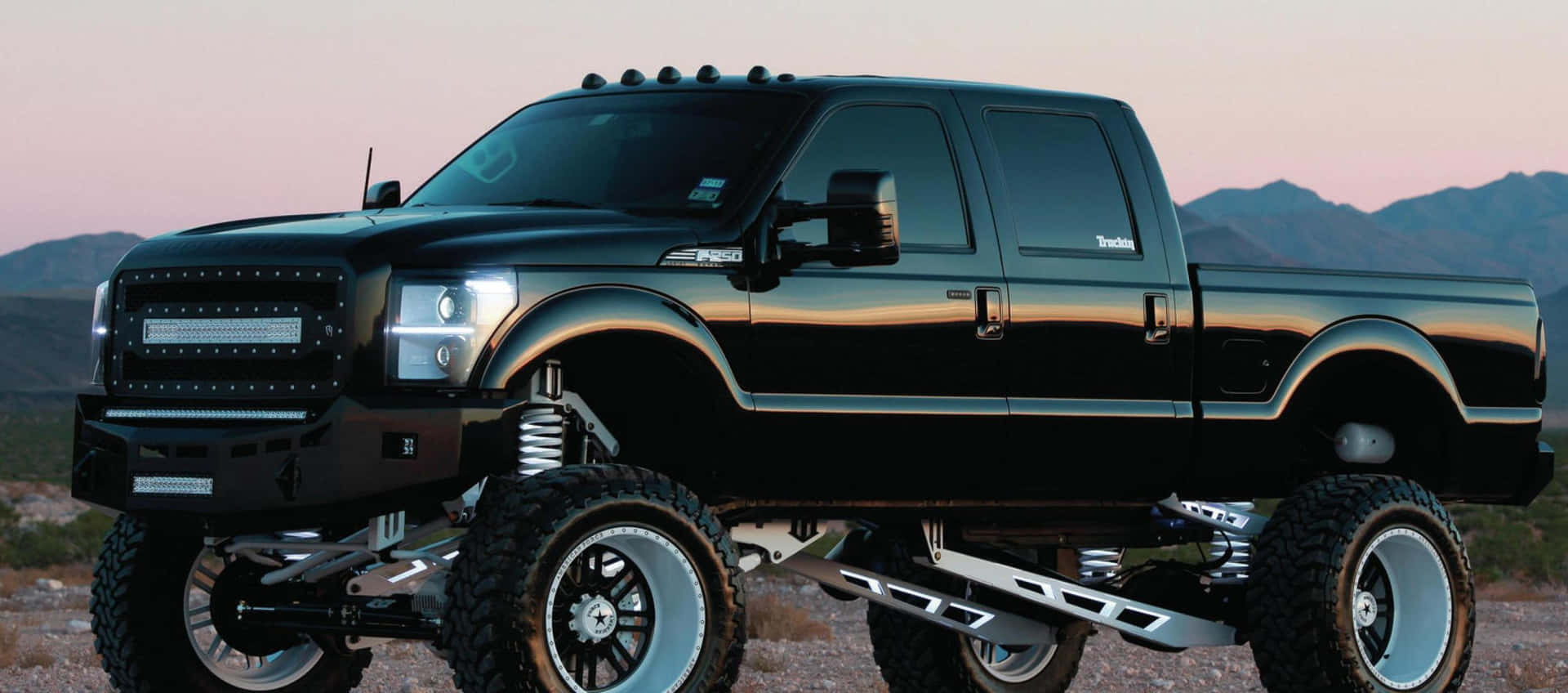 1920x848 Download A Black Truck With A Large Tire On It Wallpaper