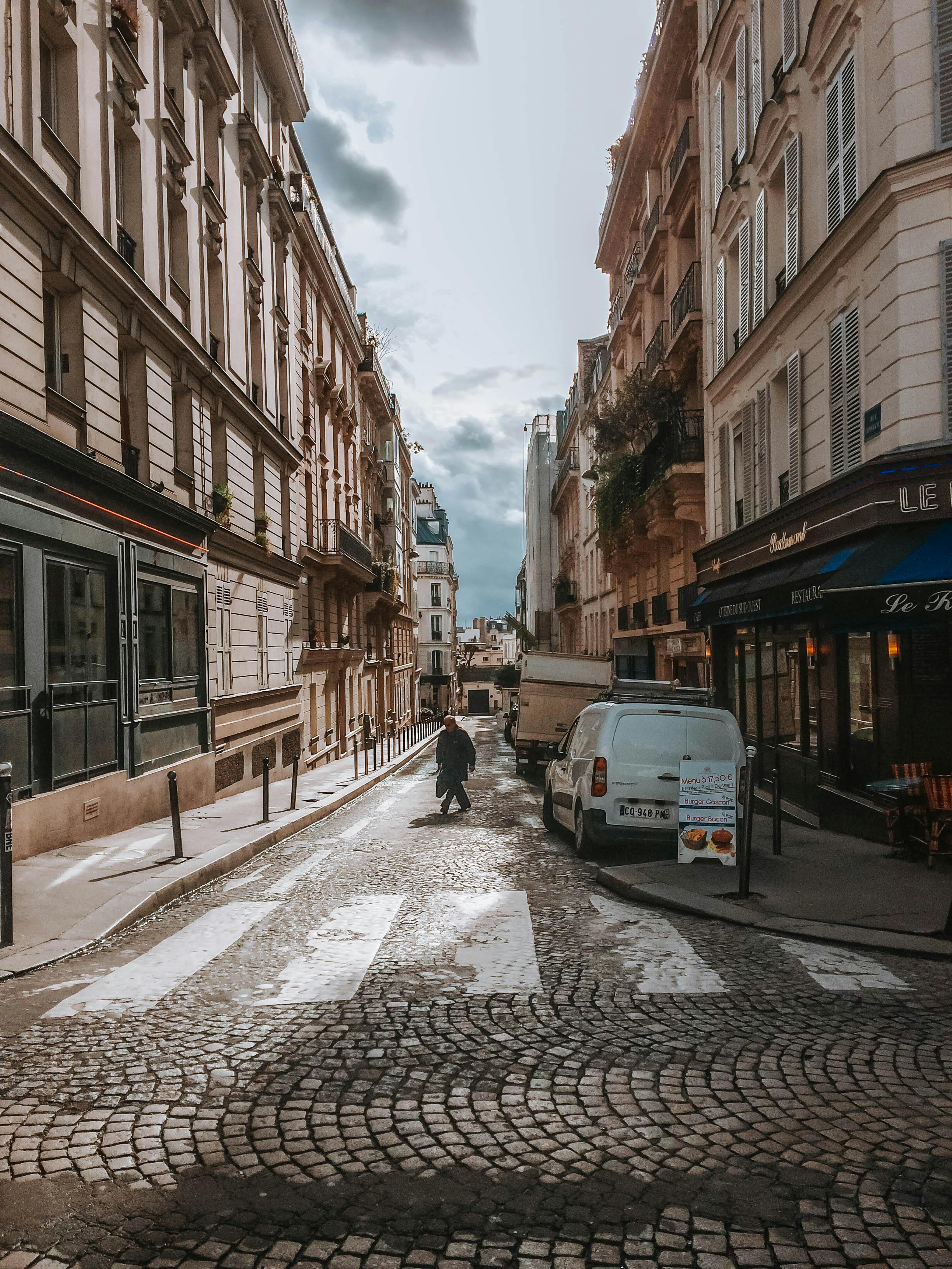 2448x3264 Person Walking On A Narrow Cobblestone Street With Parked Vehicles On The Side · Free