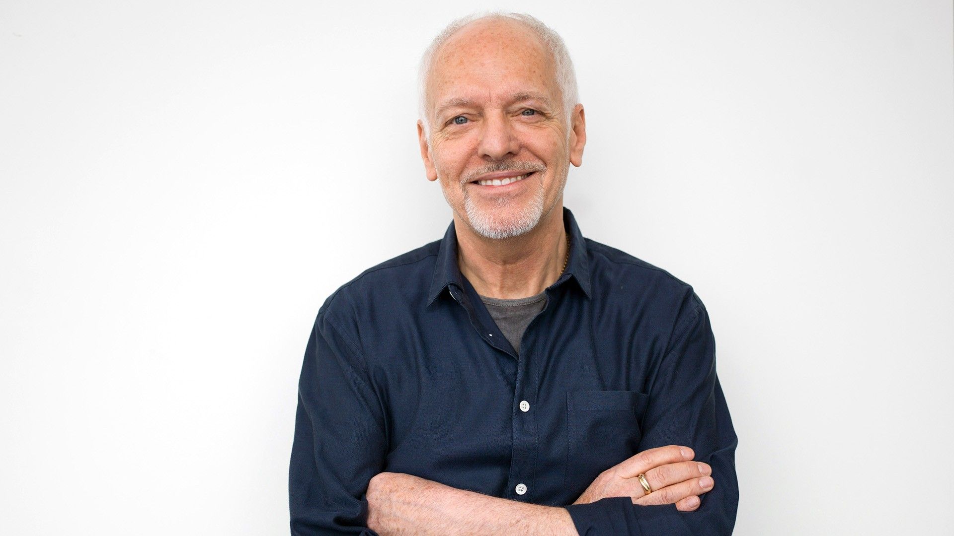 1920x1080 Smiley Peter Frampton Is Standing In White Wall Background Wearing Blue Shirt HD Peter Frampton Wallpaper