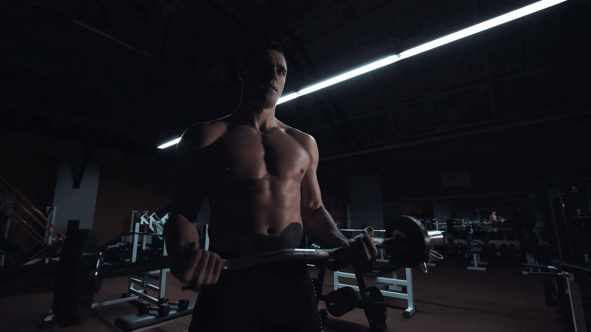 1920x1080 Muscular man doing crossfit training in a dark shadowy gym lifting weights holding a barbell at waist level in a health and fitness concept
