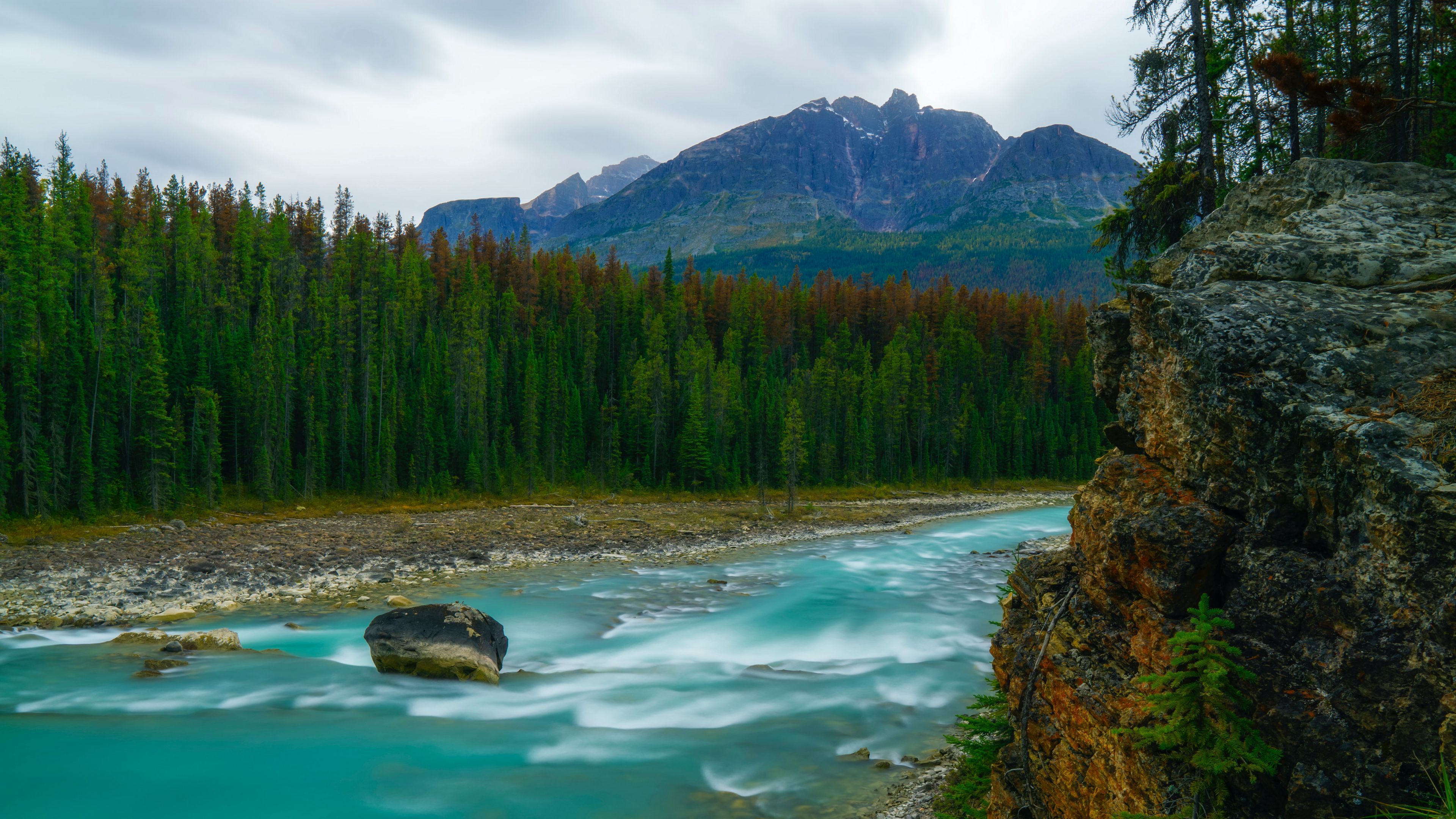 3840x2160 River Water Stream Stones Between Green Trees And Rocks In Mountains Background 4K HD Nature Wallpaper