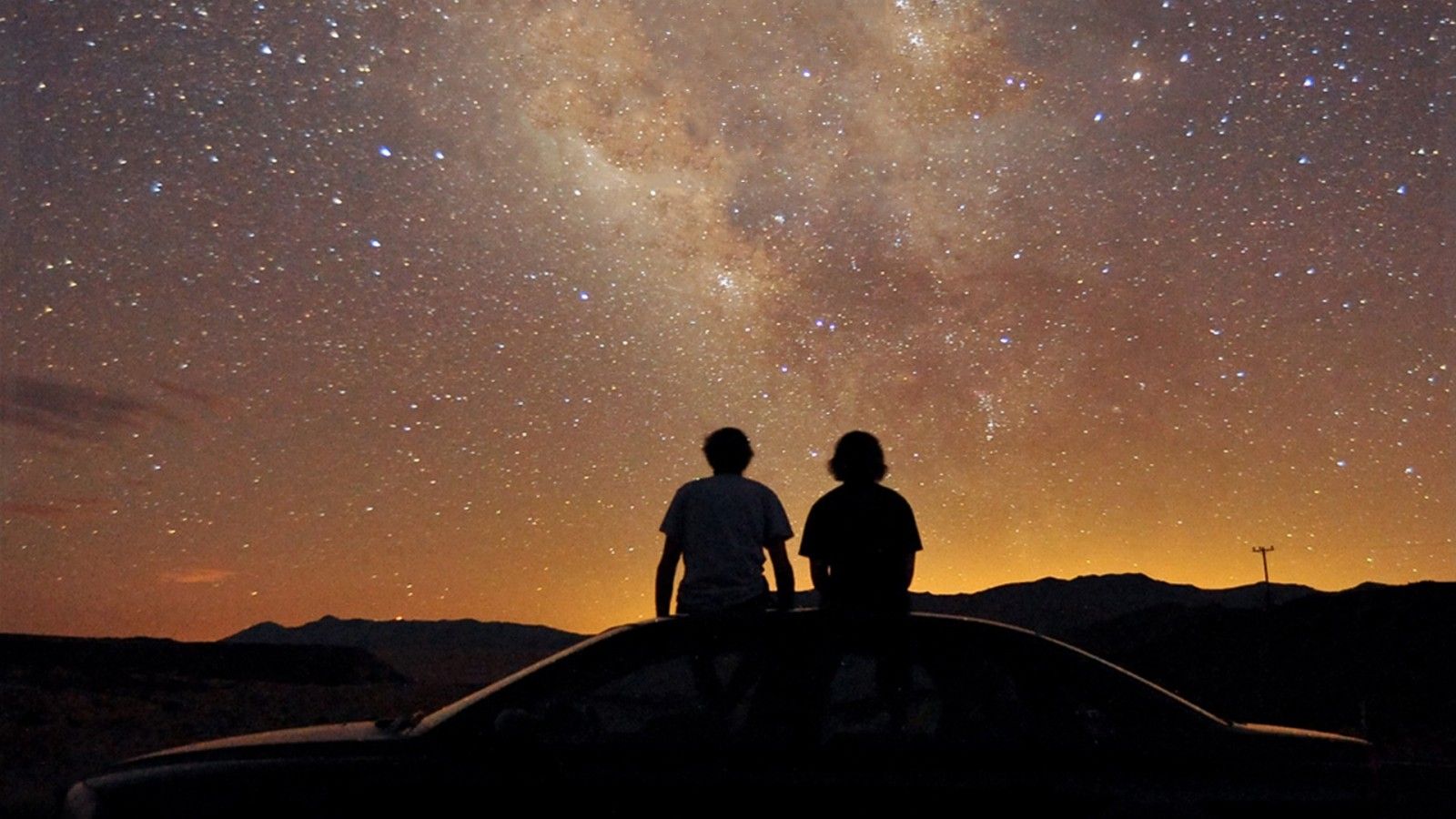 1600x900 Two Boys Sitting On Top Of Car Under Brown Sky With Stars During Sunset HD Space Wallpaper