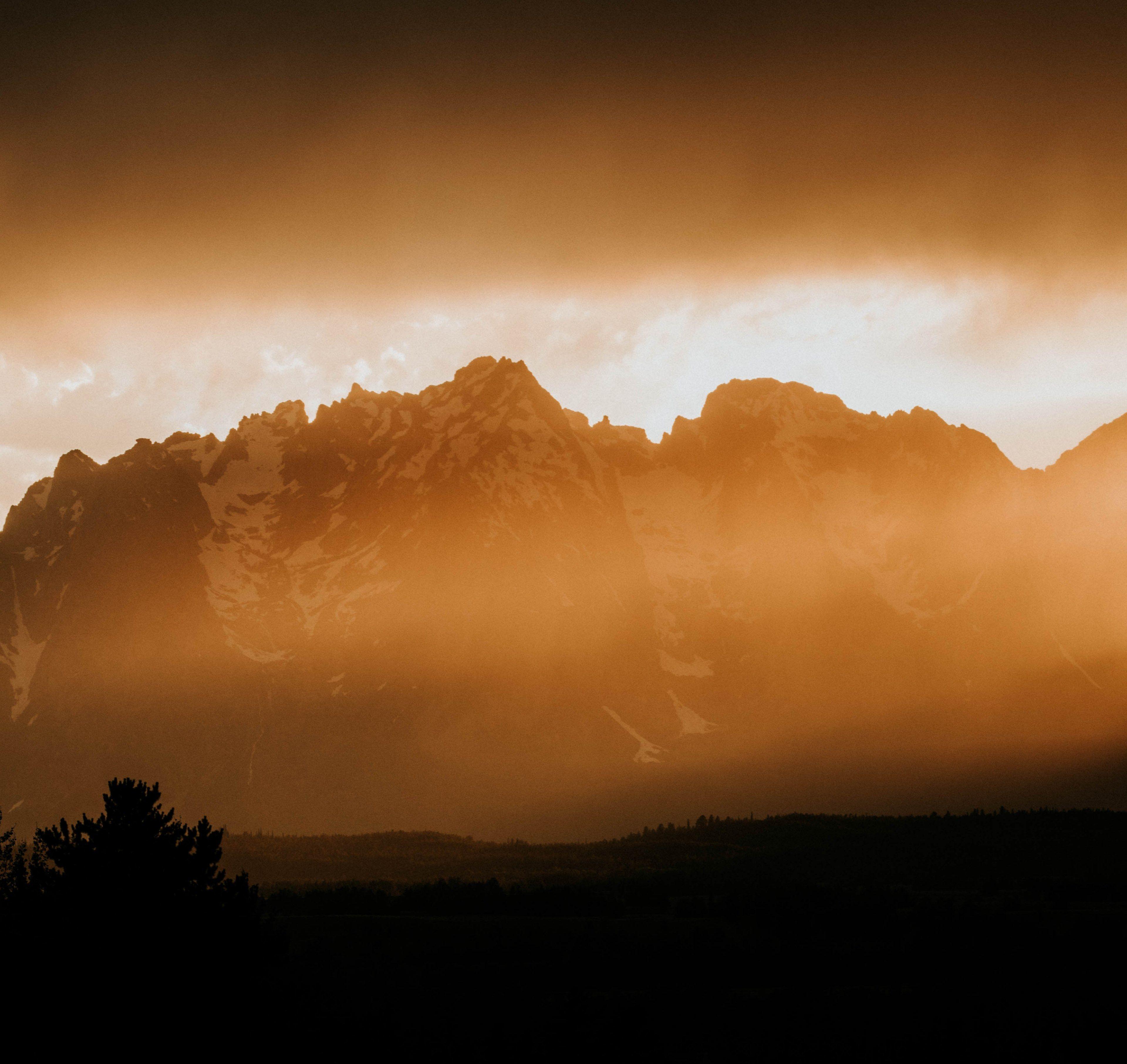 3840x3628 fog covered mountains below a cloudy sky during an orange sunset