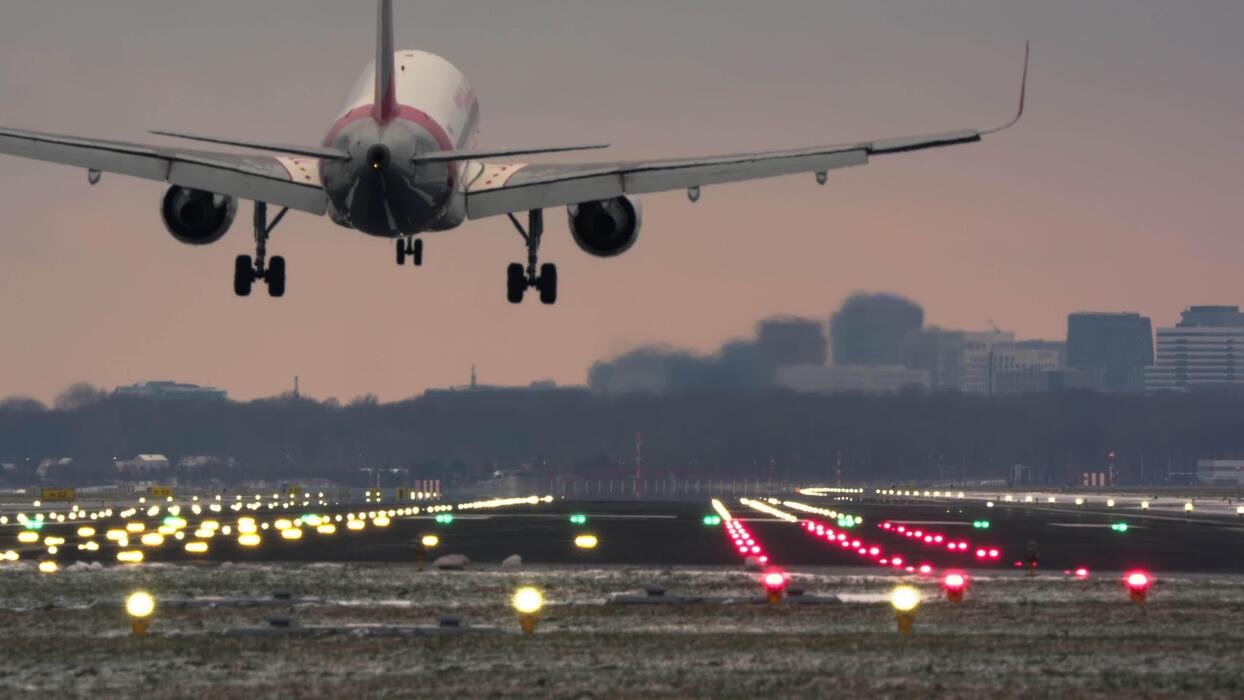 1244x700 A Large Passenger Jet Descends And Lands On A Brightly Lit Airport Runway At Dusk. The Aircraft Landing Gear Deploys As It Touches Down, With A Distant City Skyline Under A Pink Orange Sky