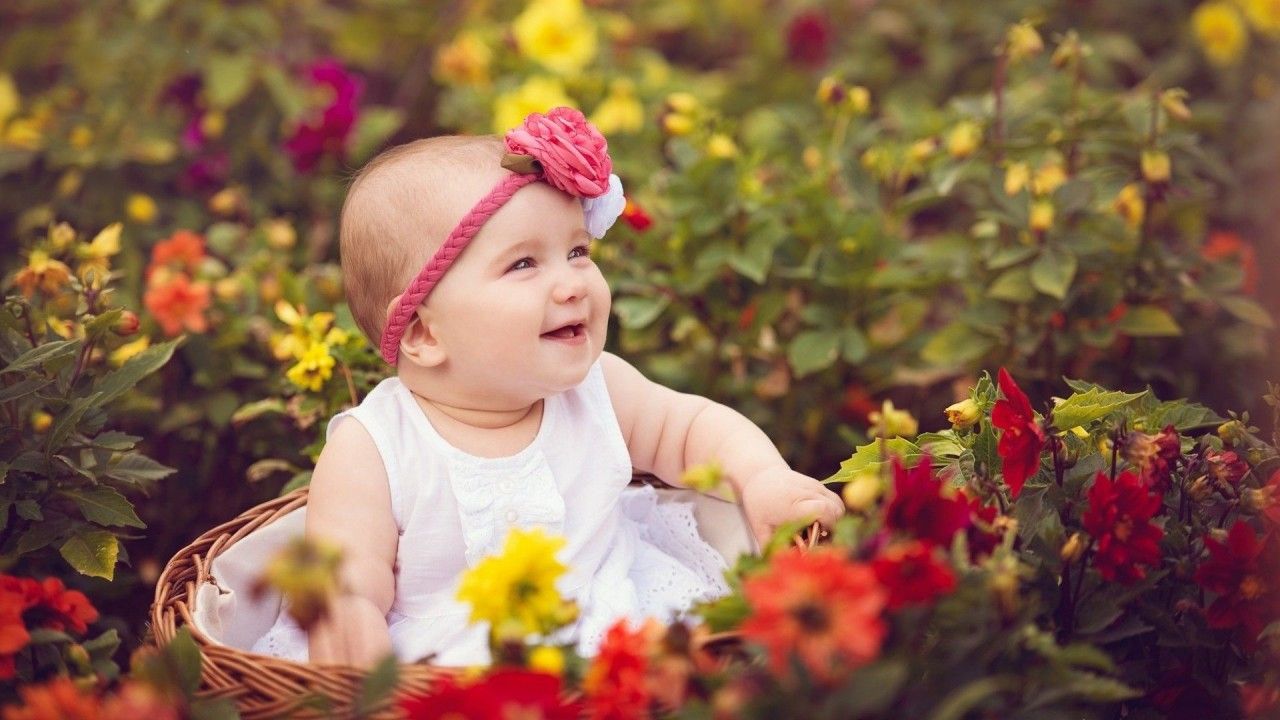 1280x720 Smiley Cute Baby Girl Is Sitting Inside Bamboo Basket Surrounded By Colorful Flowers Wearing White Dress And Flower Band On Head HD Cute Wallpaper
