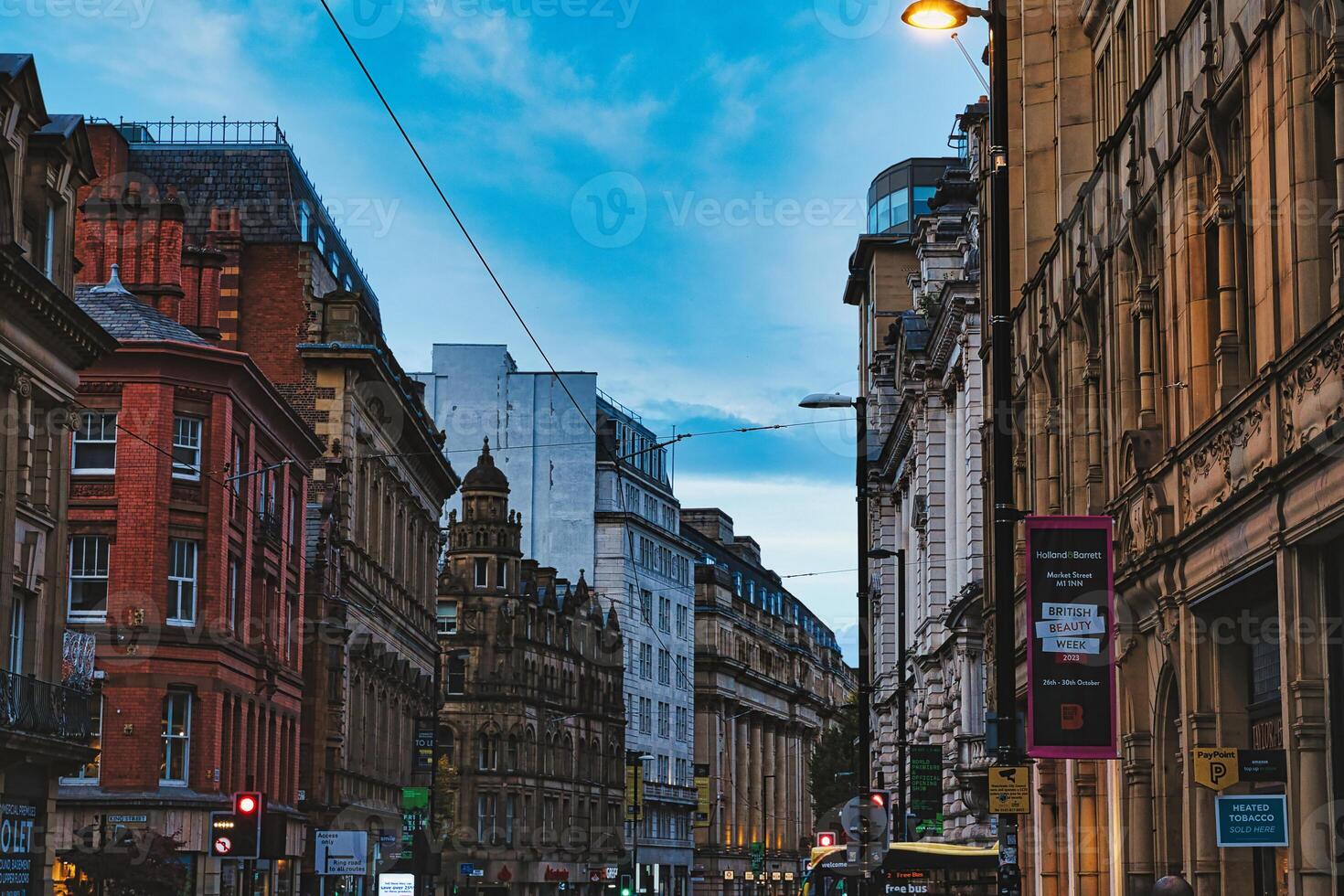 1470x980 Dusk view of a bustling city street with historic architecture and glowing street lights under a blue sky in Manchester, England