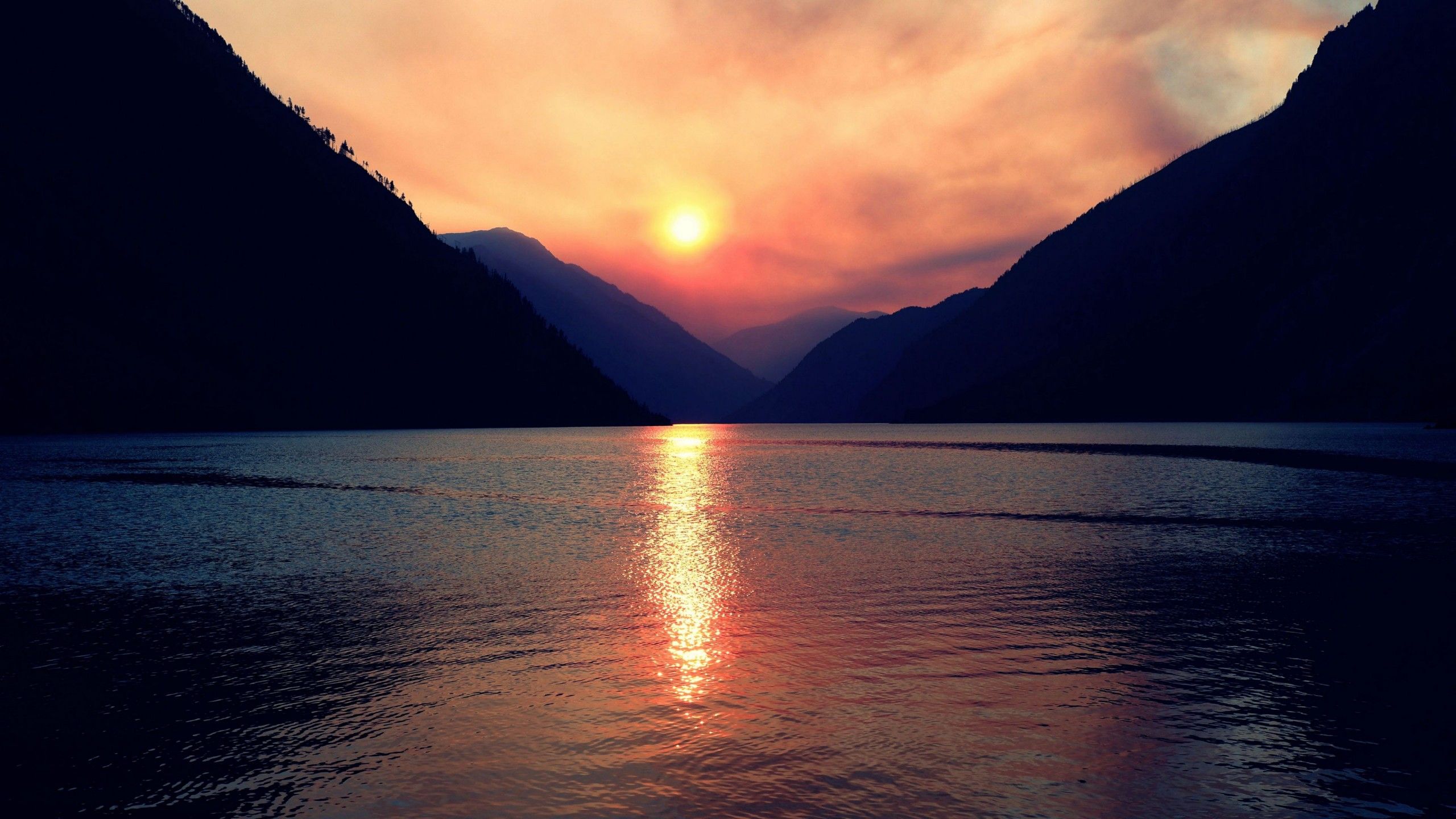 2560x1440 Mountains Lake Sun Reflection On Water In Light Red Black Clouds Sky Background 4K HD Nature Wallpaper