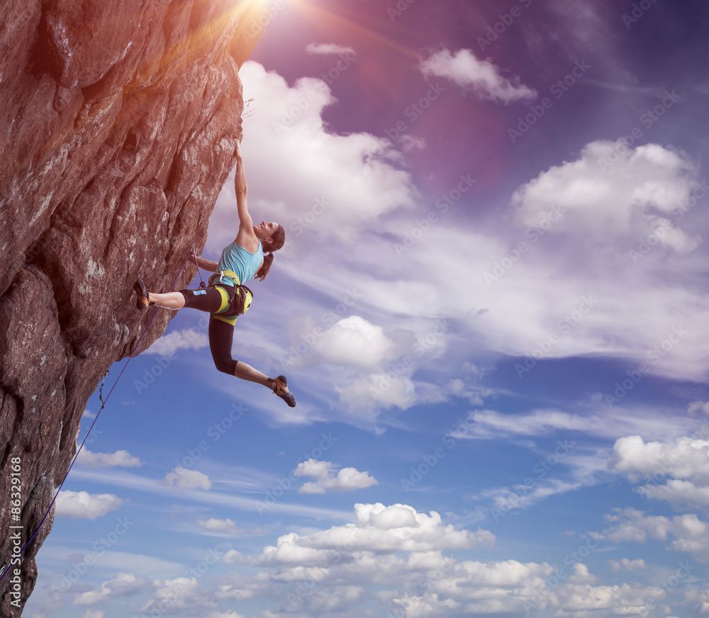 1000x872 Climber hanging on her hand. Elegant female athlete hanging at top of dangerous peak equipped with gear rope harness blue sky and terrific clouds on background and sunbeams shining from above Stock