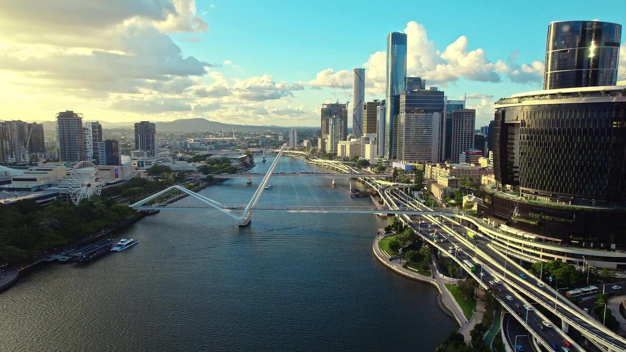1244x700 An aerial view captures Brisbane's city center with its winding river, bustling highways, towering skyscrapers, and iconic pedestrian bridges