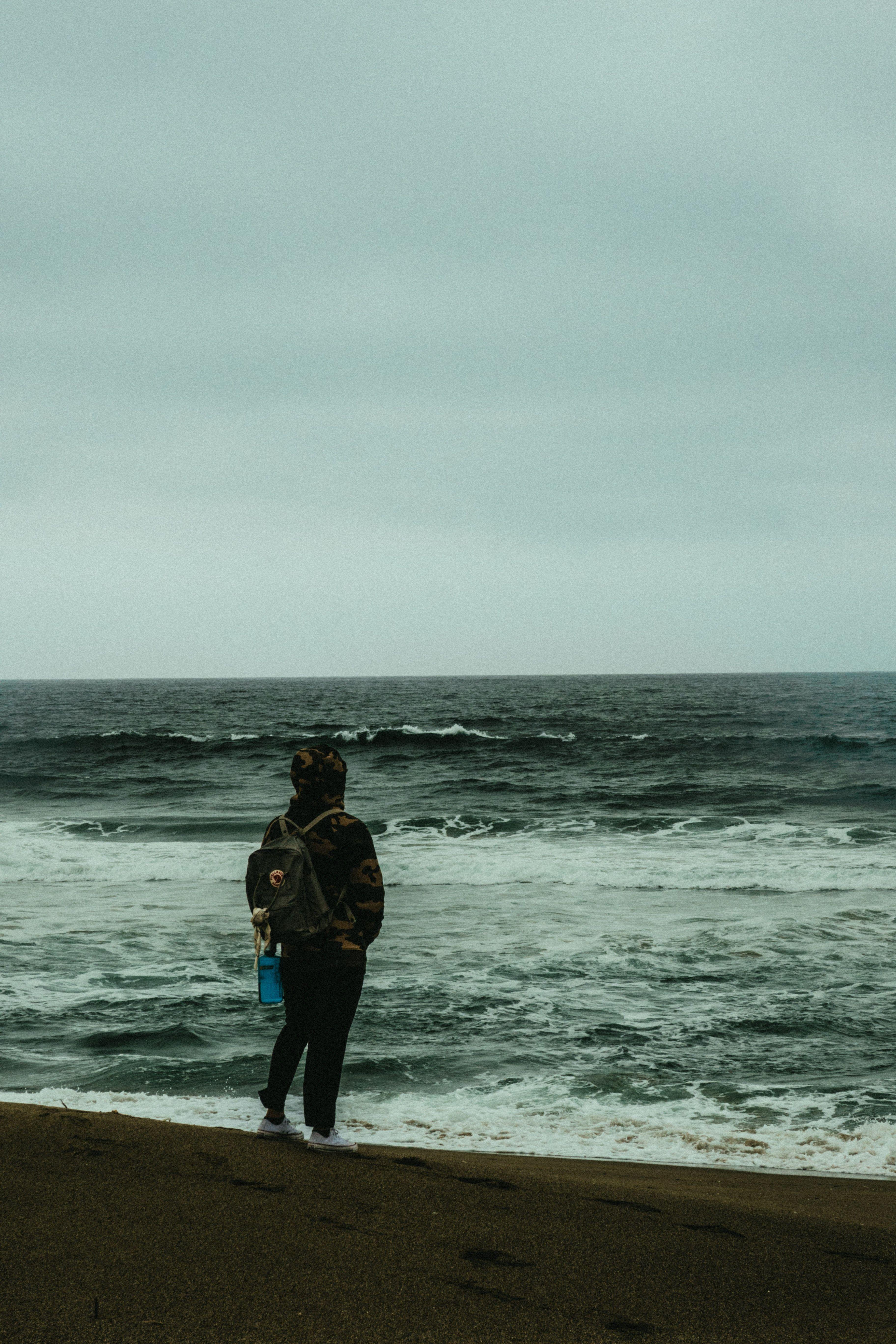 3648x5472 Photo of Person Carrying Backpack Standing Alone on the Beach Overlooking the Horizon · Free