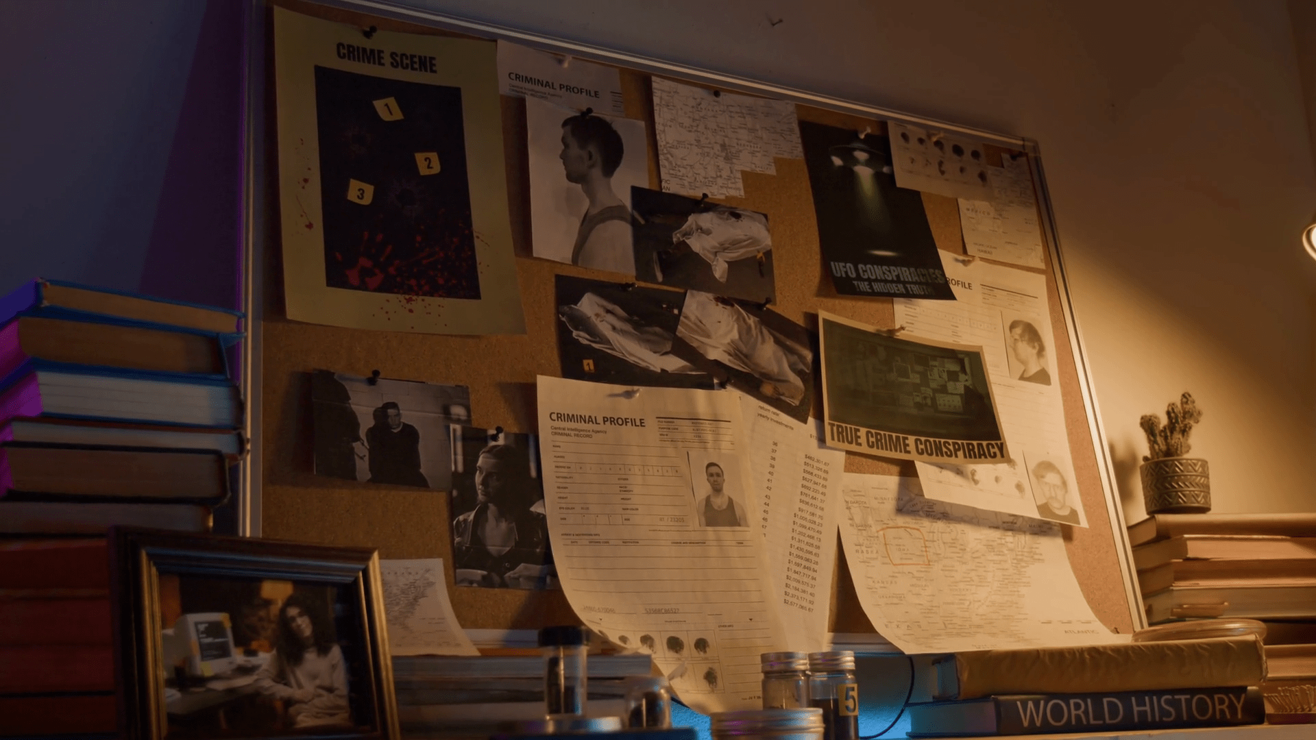 1920x1080 No people view of notice board with picture and prints pinned to it including crime scene photo, criminal profiles and maps of areas on shelf near framed photograph, books and glass bottles