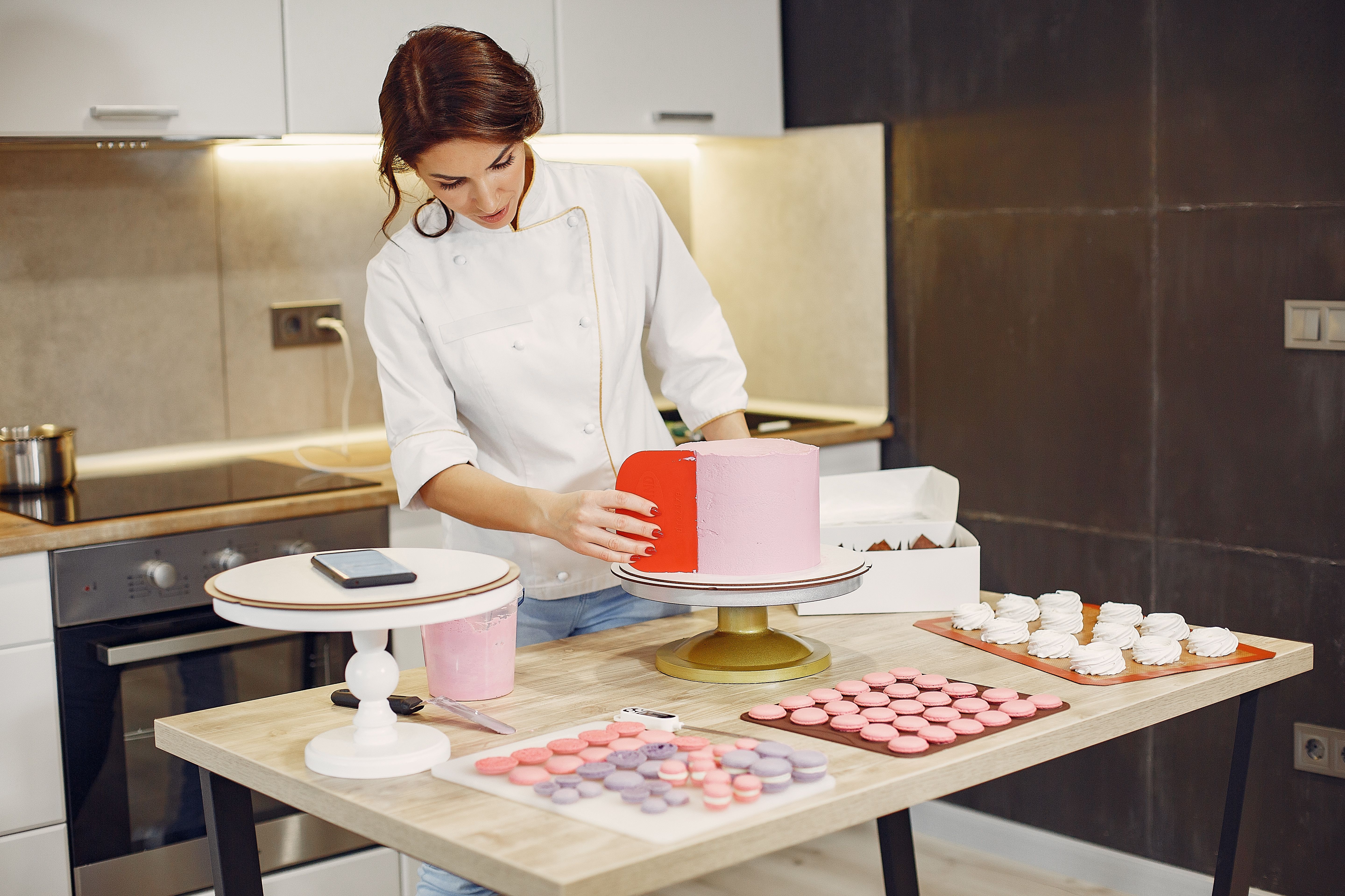 5760x3840 Focused female pastry chef preparing cake and desserts with smartphone aside in modern kitchen · Free