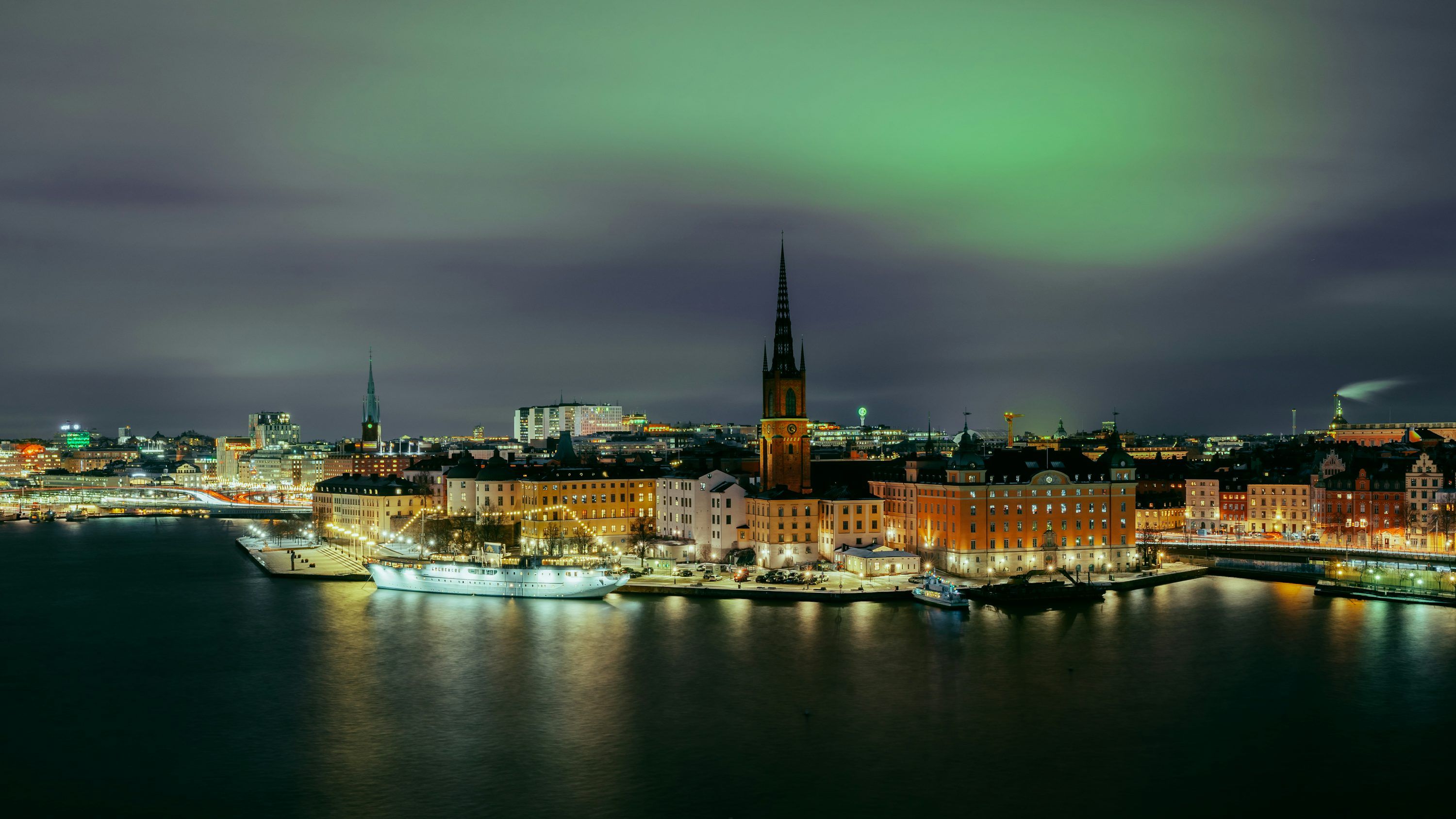 3000x1688 A view of a city at night with a green light in the sky