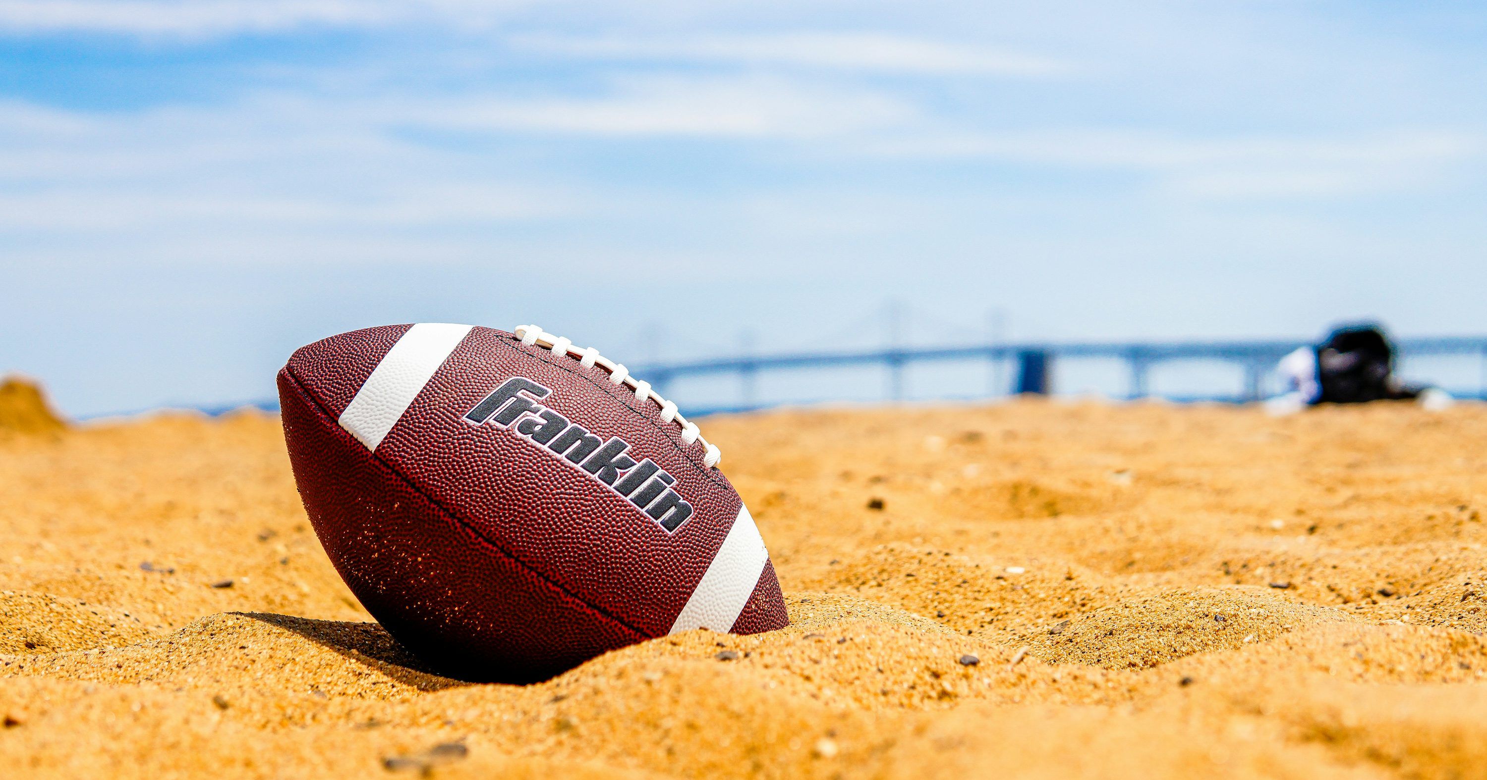 3000x1575 American football partially buried in sand on a sunny beach