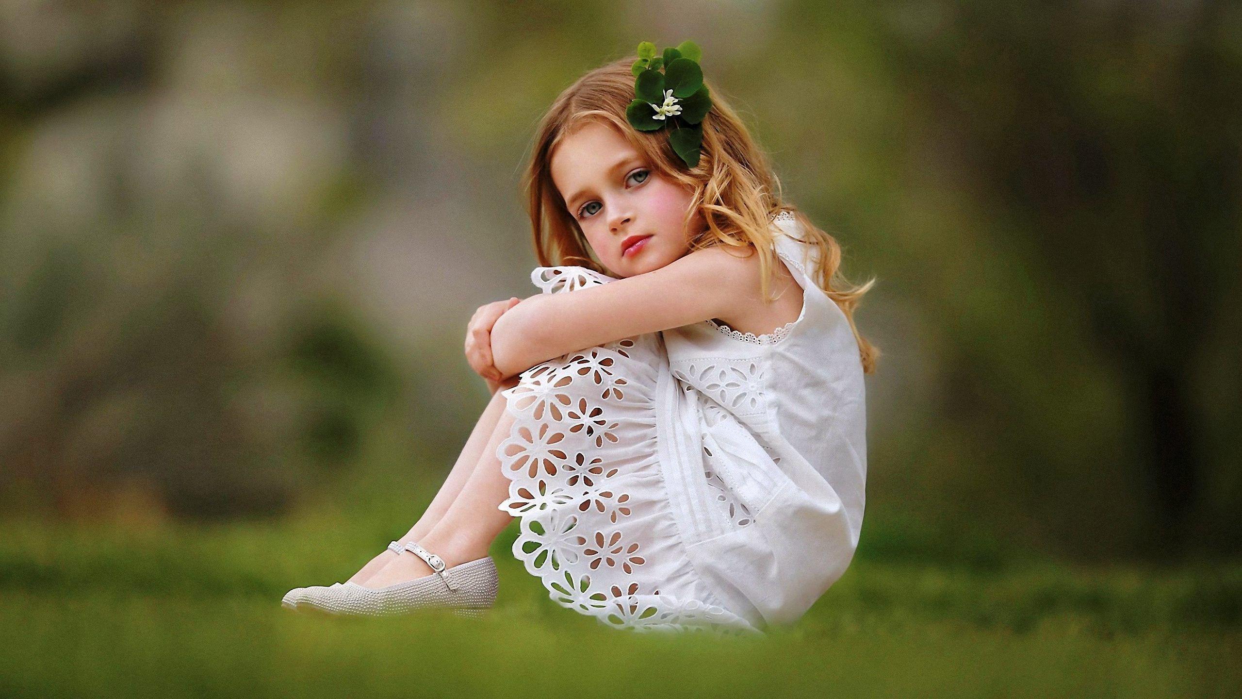 2560x1440 Cute Girl Baby Is Wearing White Dress Sitting On Grass Facing One Side And Having Green Leaves On Head In A Blur Background HD Cute HD Wallpaper