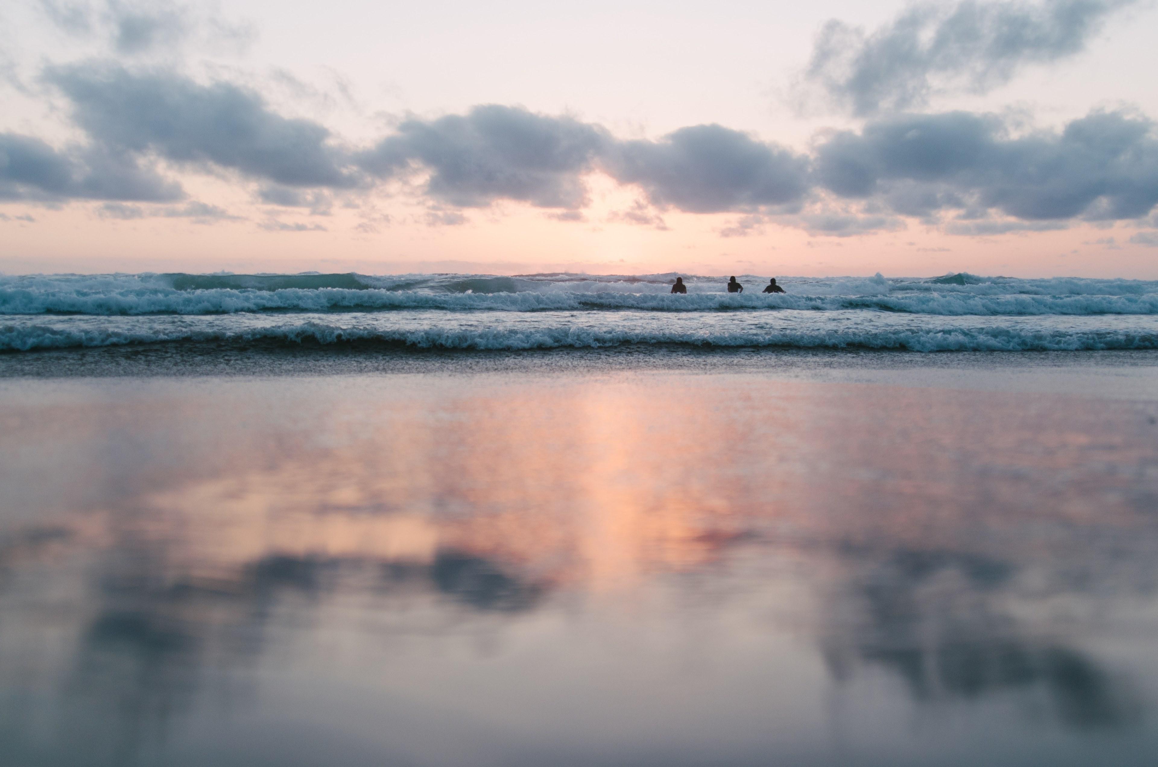 3840x2543 Wallpaper / three people in ocean waves with a blurred reflection of a partly cloudy pastel sunset in the foreground, cloud ocean sunset and beach HD 4k wallpaper