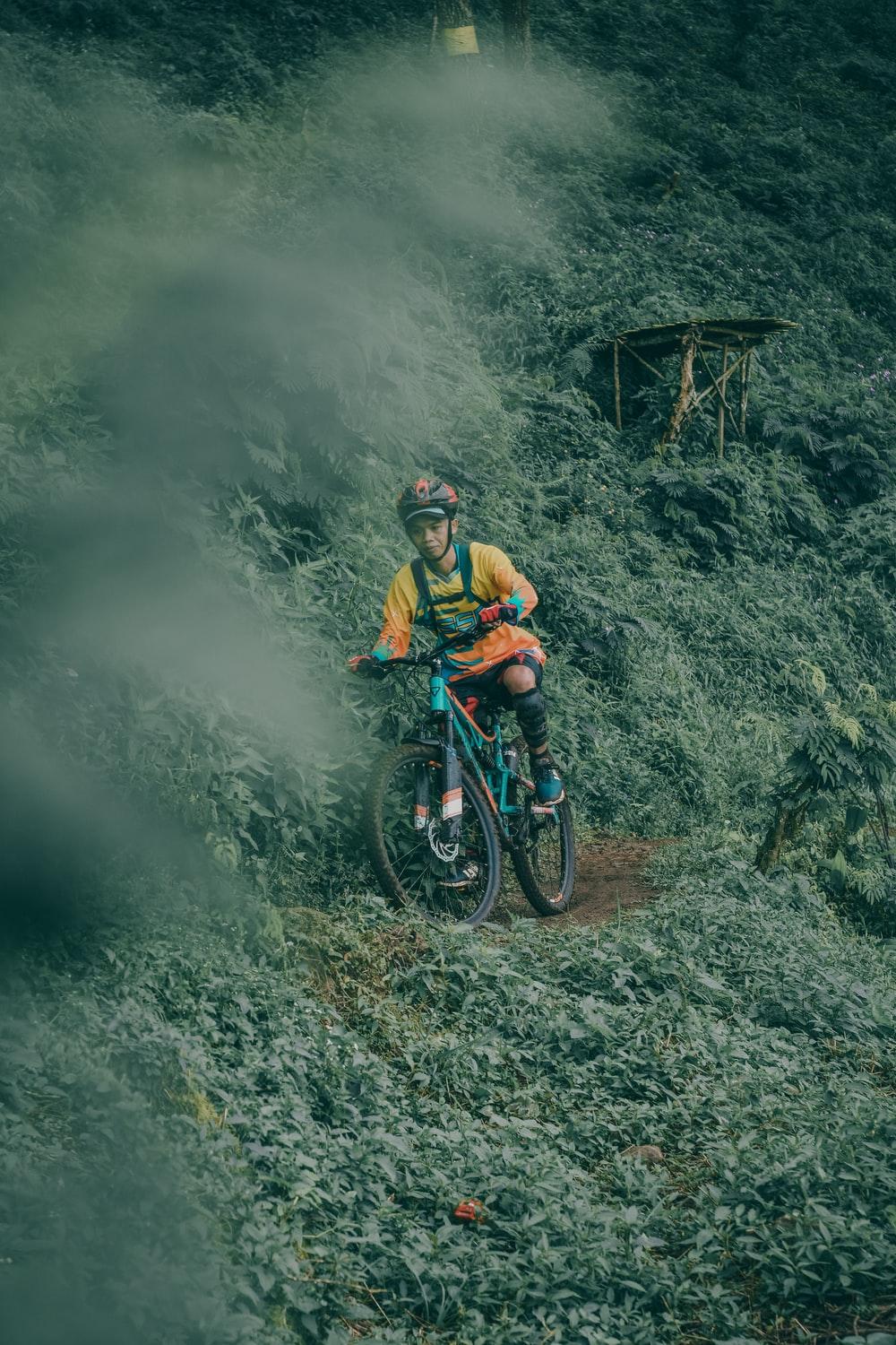 1000x1500 Man Riding Teal Full Suspension Mountain Bike On Edge Surrounded By Green Leaf Plants Photo