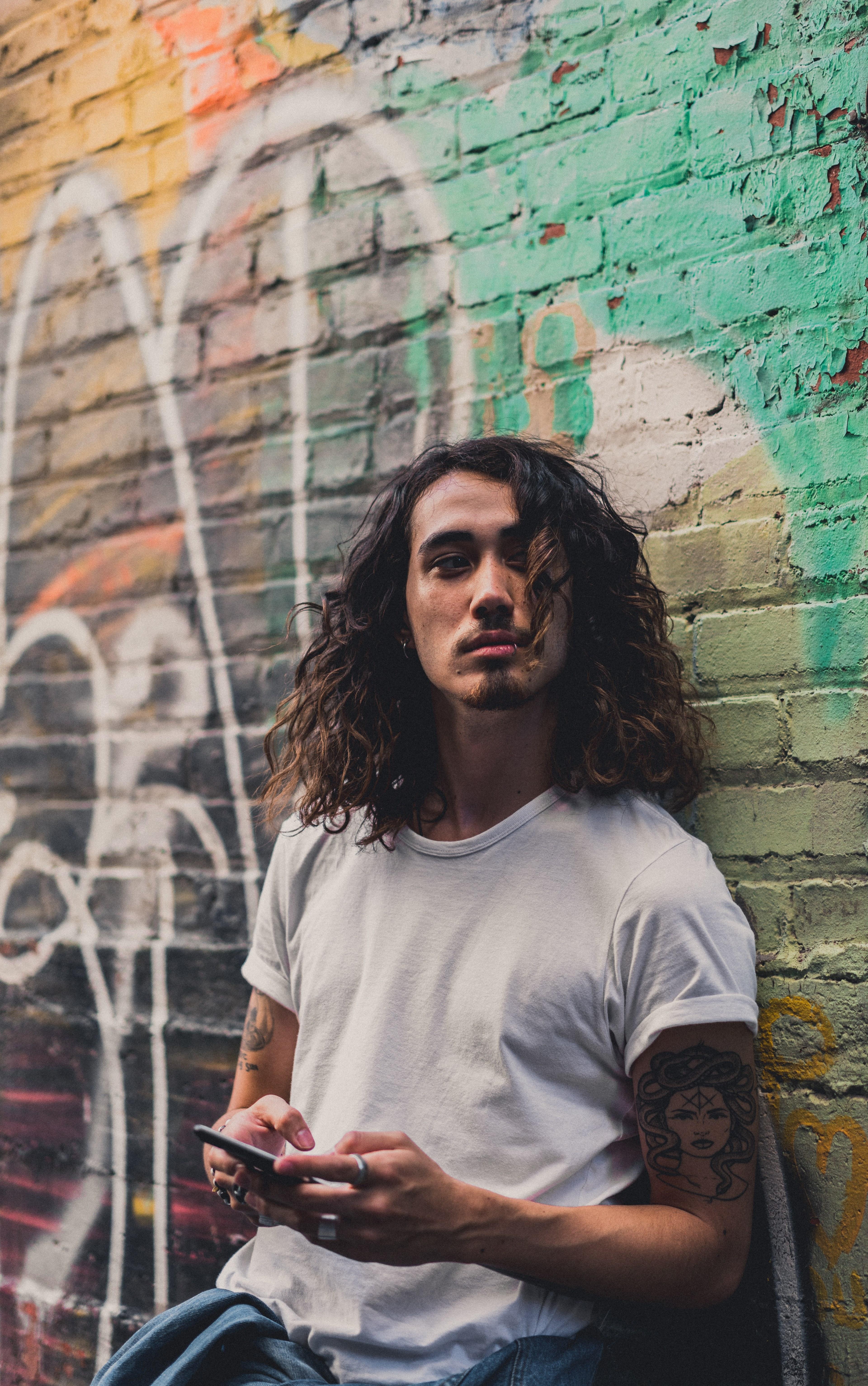3840x6128 Wallpaper / a man with long curly hair leans against a graffitied wall in montreal using an iphone, whos there 4k wallpaper free download